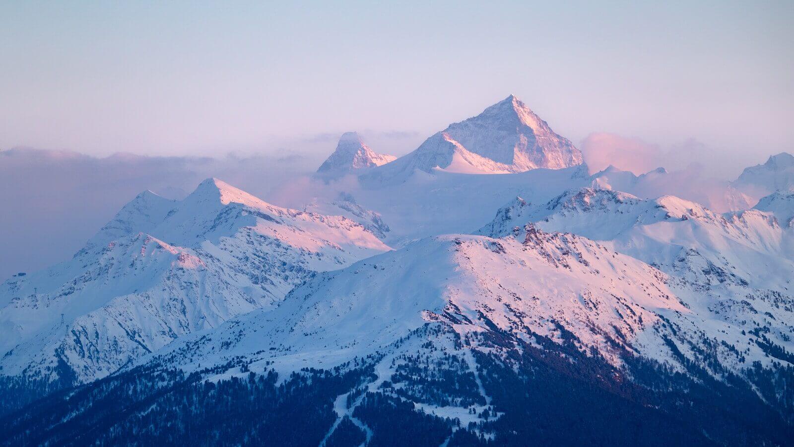 Snow-covered mountains bathed in soft pink sunrise light.