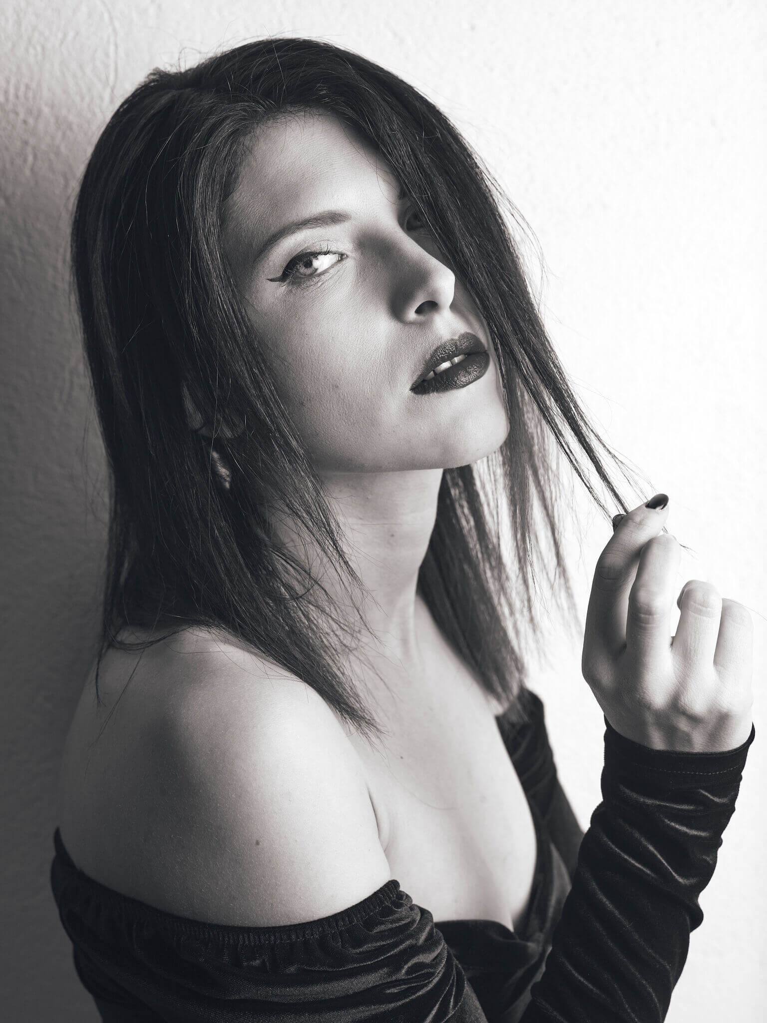 A woman with dark hair and makeup poses against a plain wall while holding a strand of her hair.